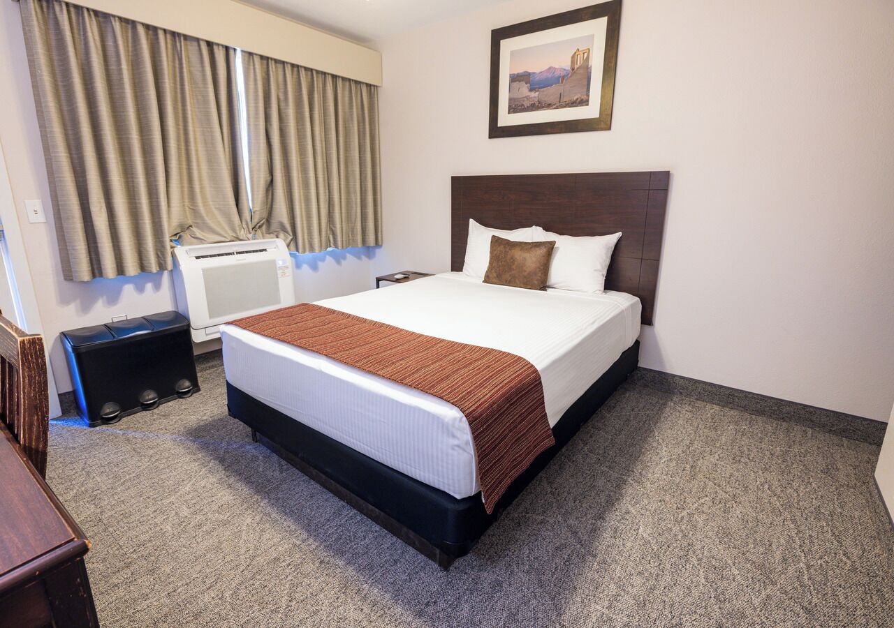 A hotel room with a large bed, a headboard, a window with curtains, a wall-mounted air conditioner, and a framed picture above the bed.