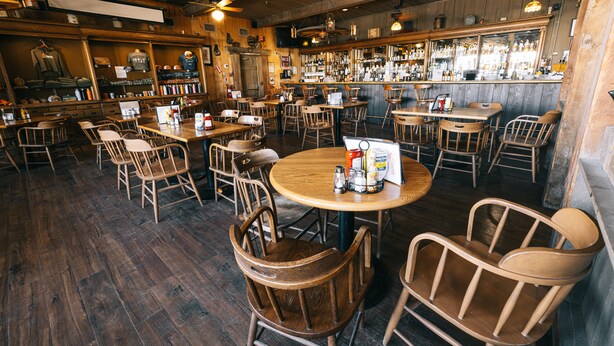 Un interior de restaurante con mesas y sillas de madera, una zona de bar con botellas y una decoración rústica.