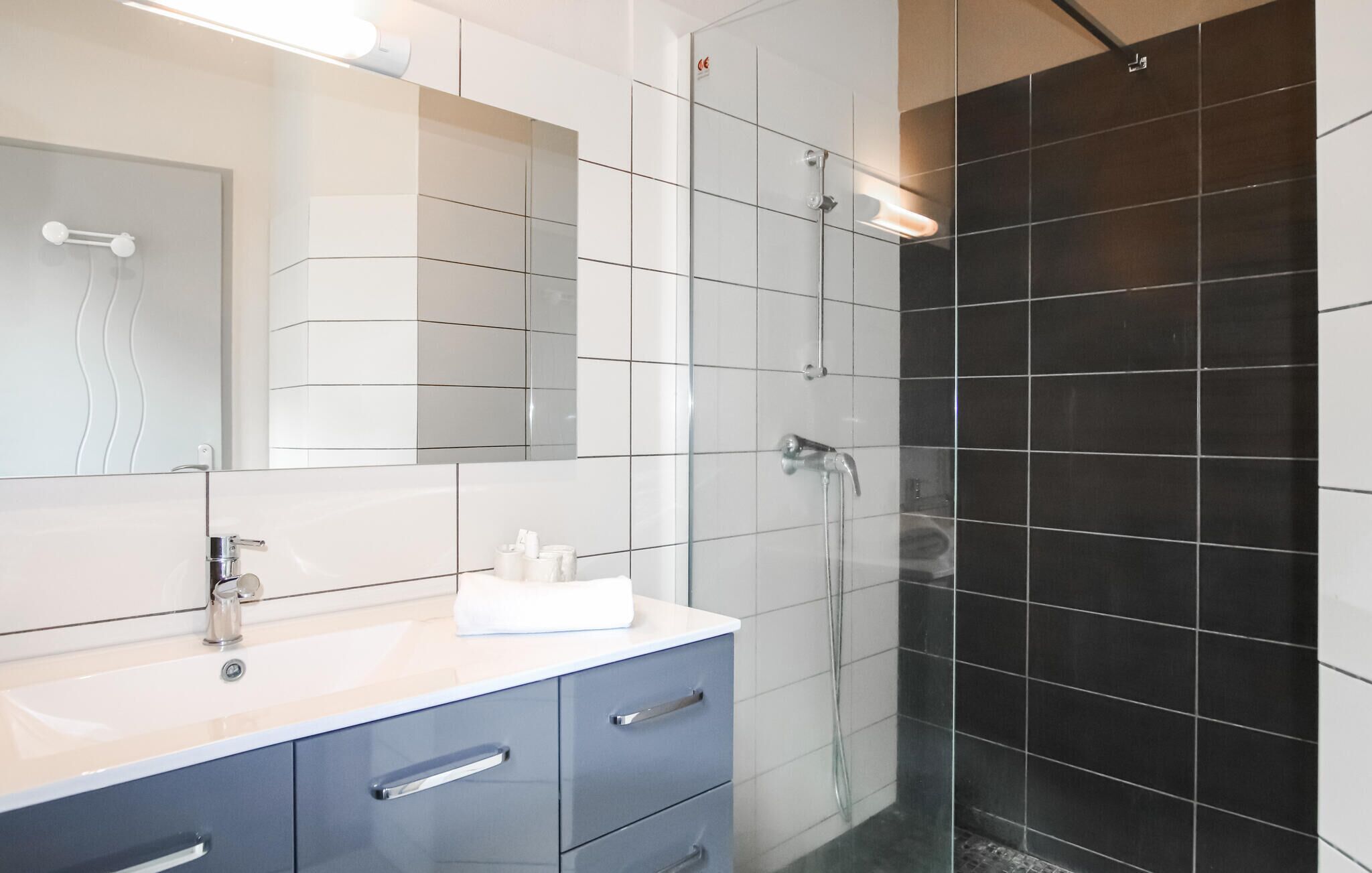 Ein modernes Badezimmer mit einem weißen Waschtisch, einem großen Spiegel und einem Duschbereich mit schwarzen Fliesen.