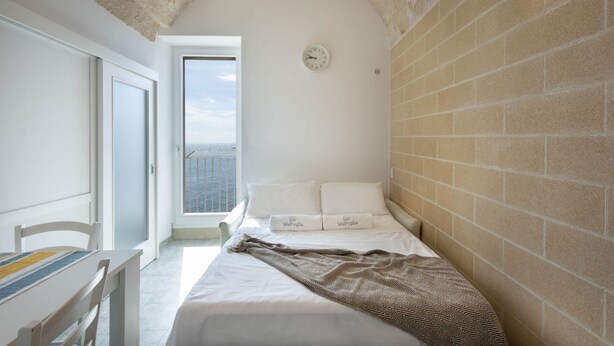 A bedroom with a bed, a clock on the wall, and a view of the sea through a window.