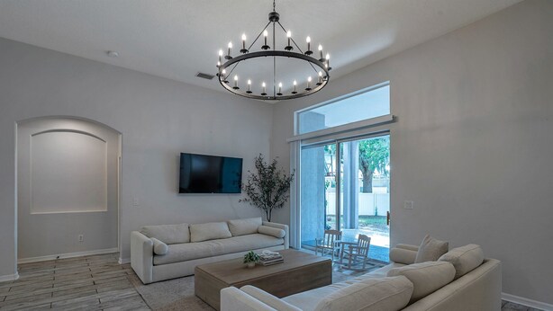 A modern living room with a large chandelier, a flat-screen TV mounted on the wall, and a large sliding glass door leading to an outdoor area. - Large 5 bedroom 3 full bath house close to beaches, shopping  and parks. (Parrish)