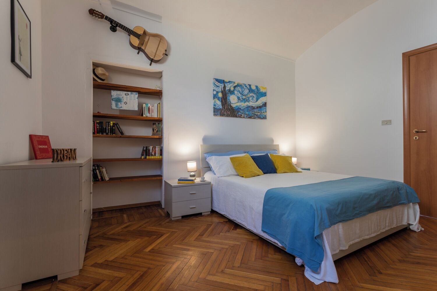 Une chambre à coucher avec un lit, une table de chevet, une bibliothèque, une guitare et un tableau accroché au mur.