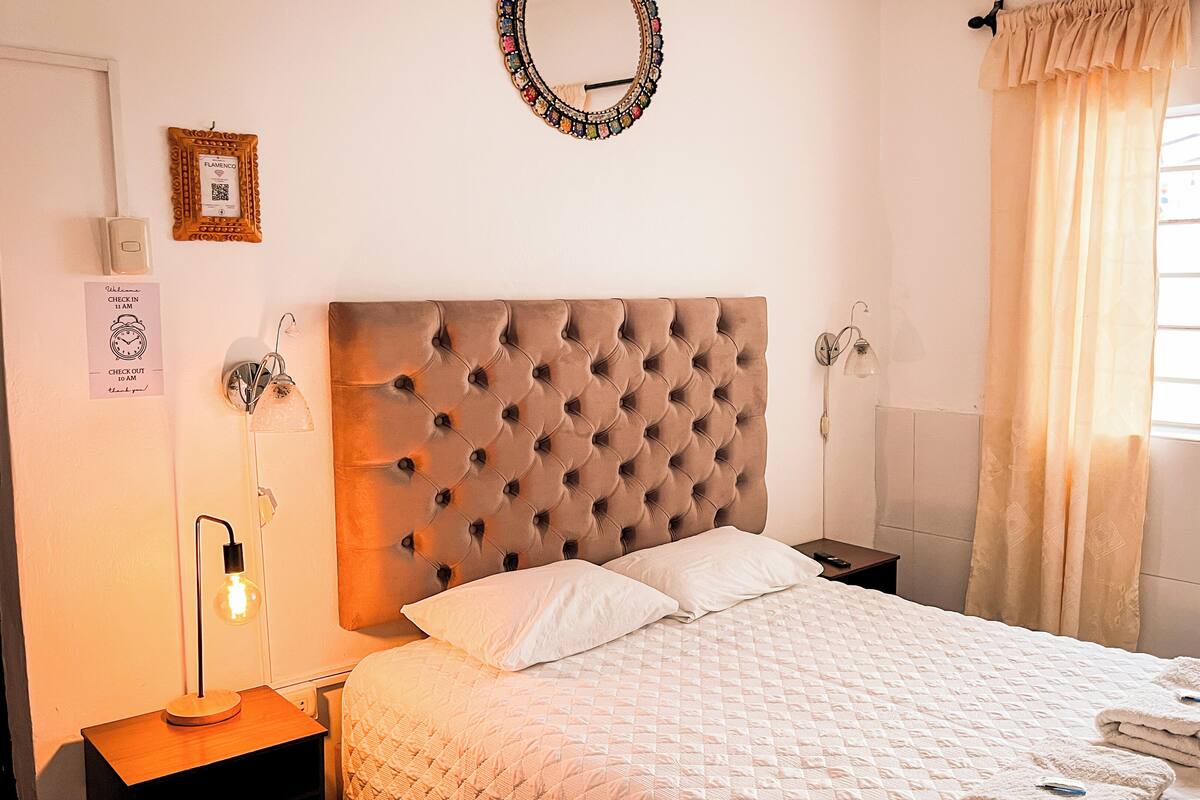 Ein Schlafzimmer mit einer gepolsterten Kopfteile, einem Bett mit weißer Bettwäsche, einem Nachttisch mit Lampe, einer Wanduhr und einem Fenster mit Vorhängen.