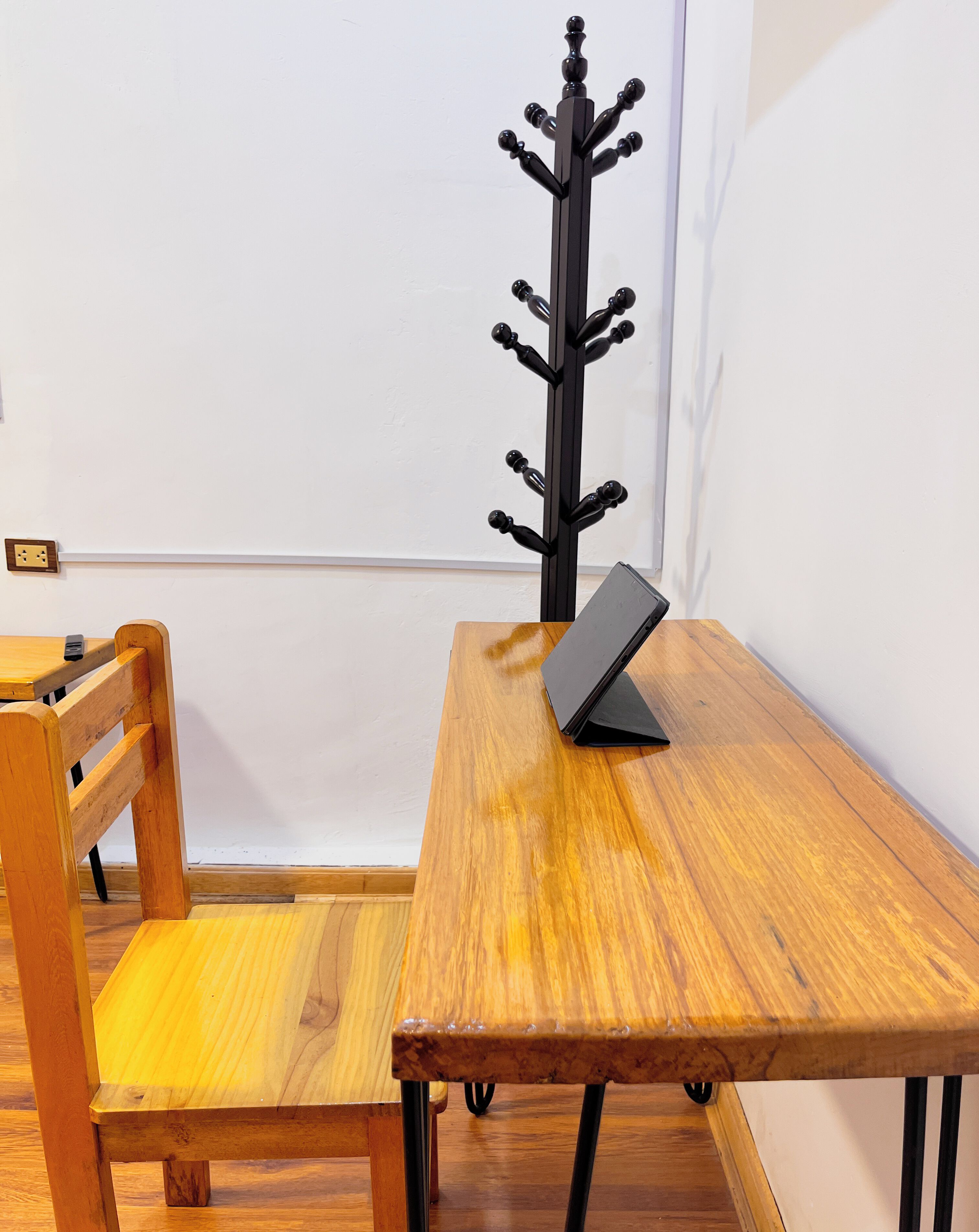 A wooden dining table with a black electronic device on it, a wooden chair, and a black coat rack against a white wall.