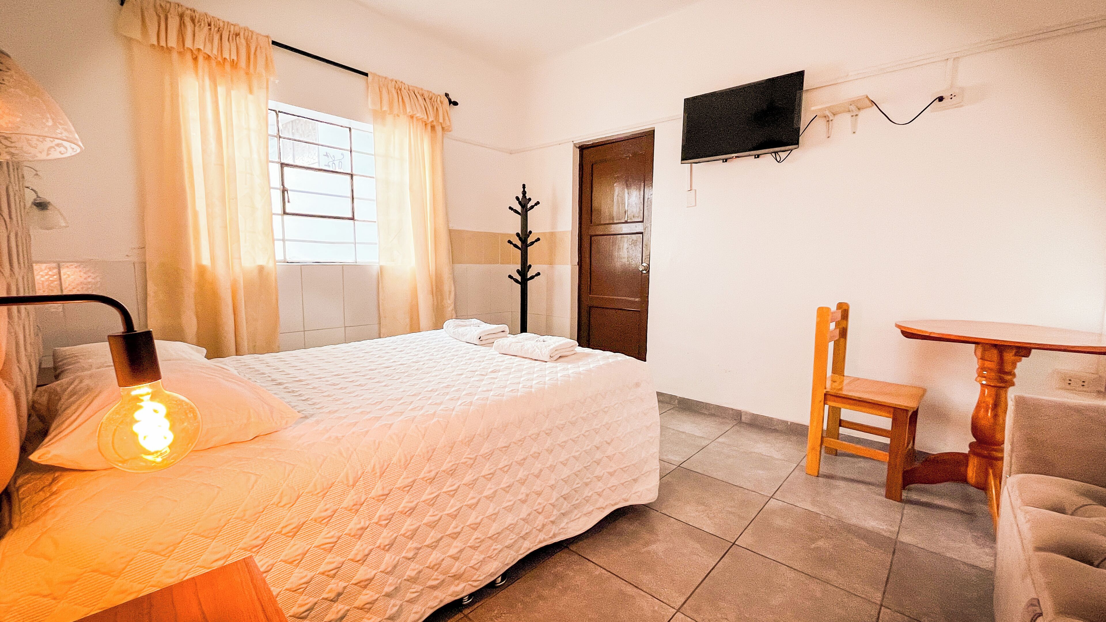A hotel room with a bed, a chair, a small table, a TV mounted on the wall, and a window with curtains.