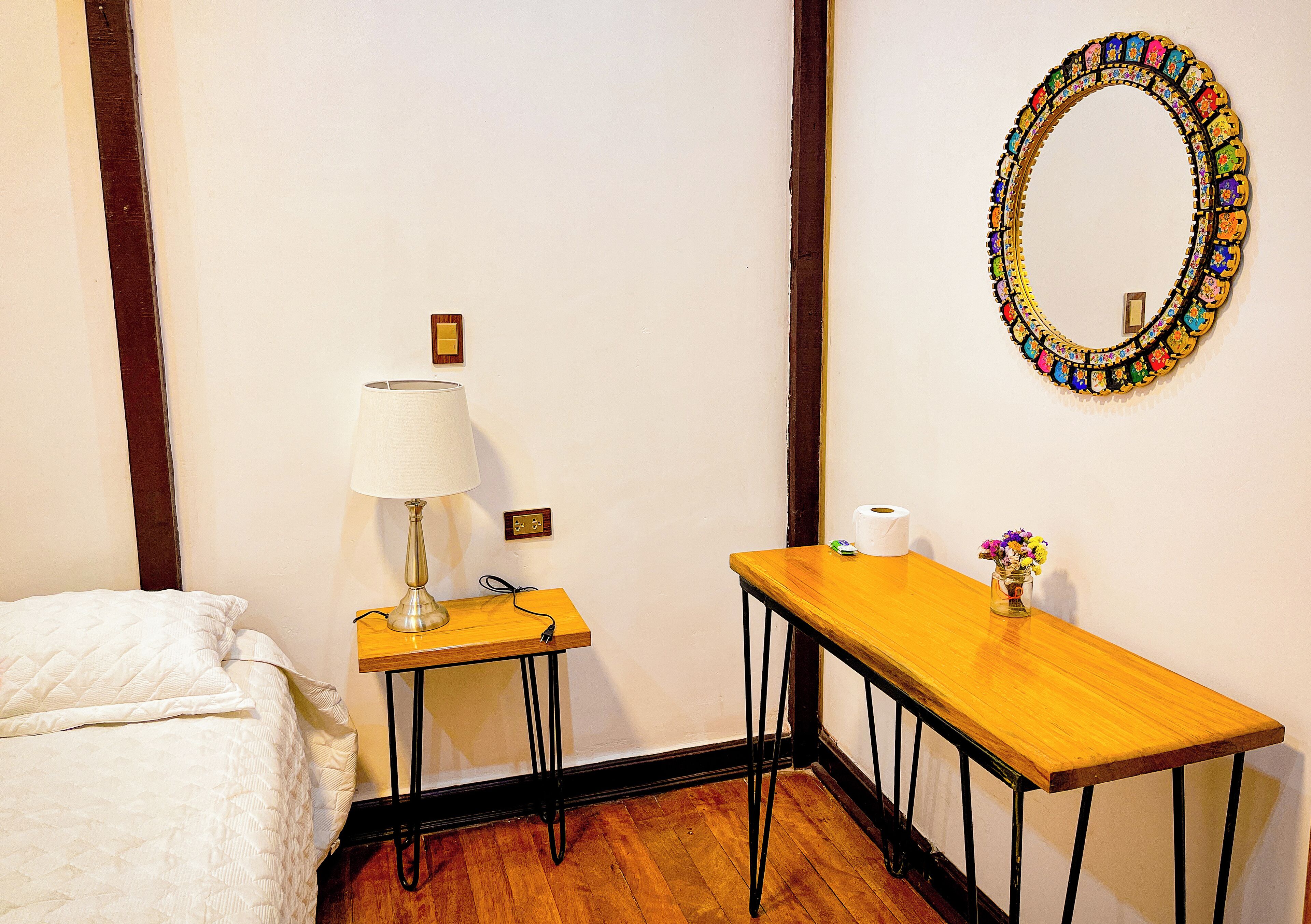A bedroom with a bed, a bedside table with a lamp, a mirror, and a small table with a vase of flowers.
