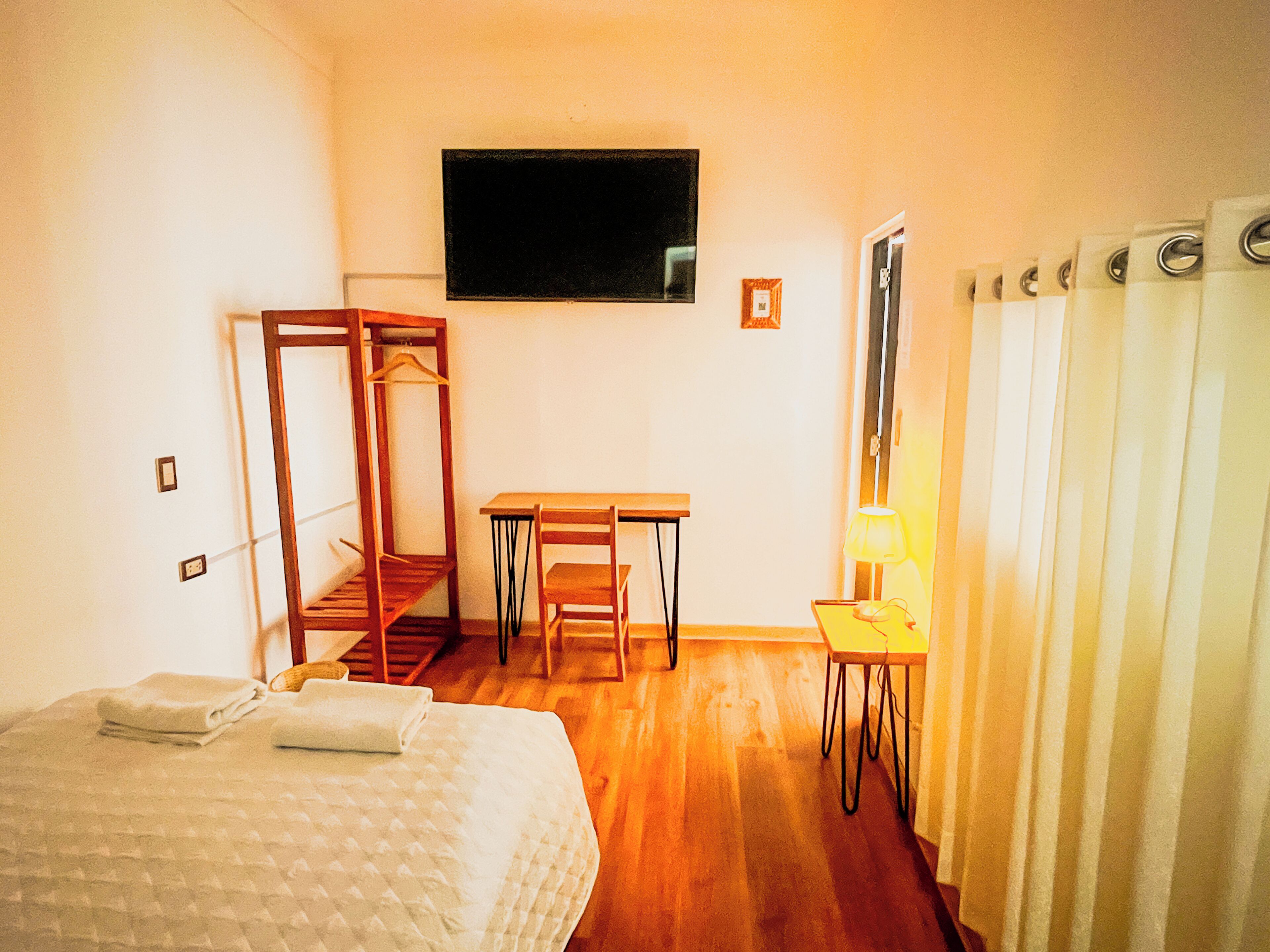 A room with a bed, a desk, a chair, a hanging coat rack, a small table with a lamp, and a window with a black frame.
