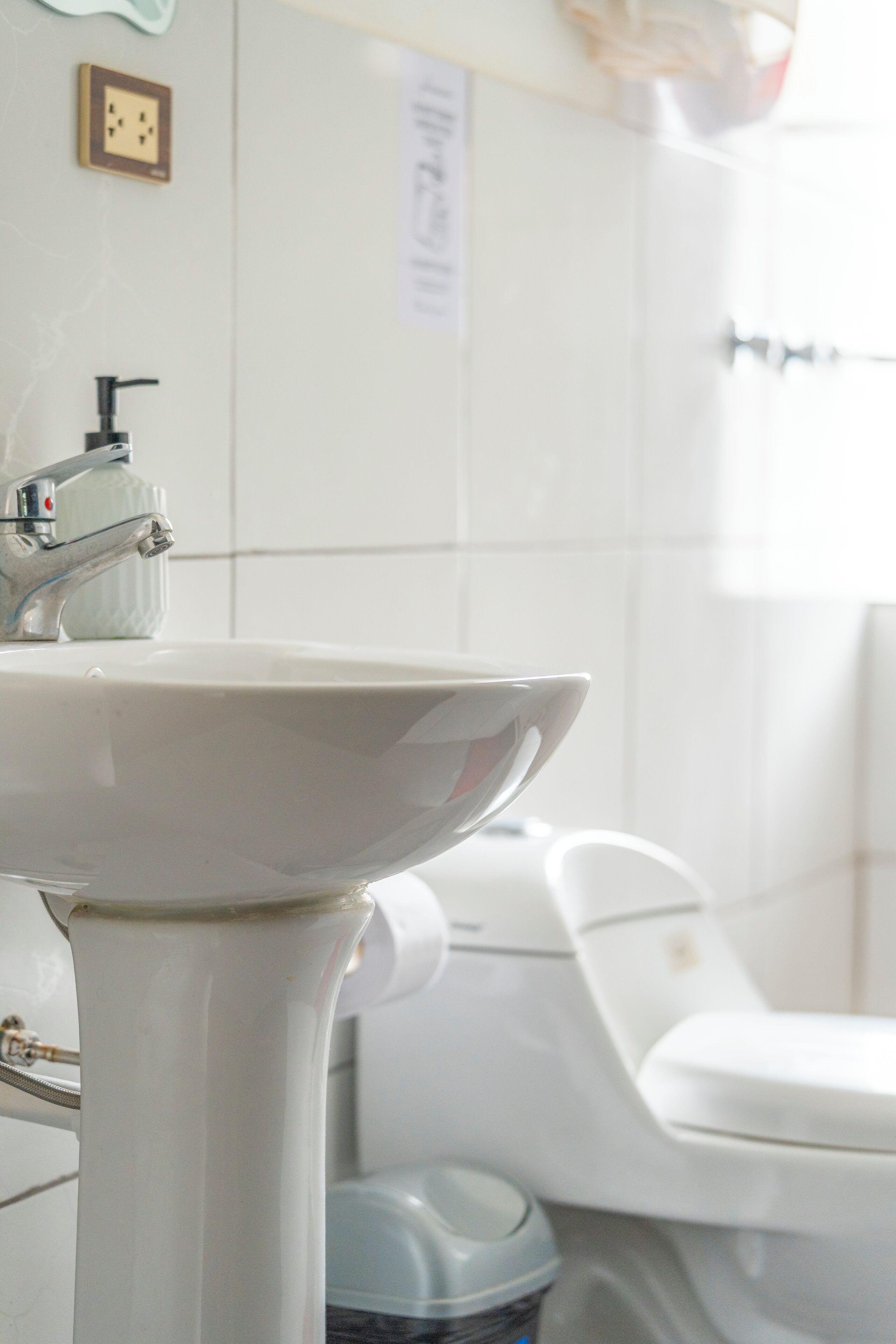 Une salle de bain avec un lavabo, un robinet et des toilettes blancs. On y trouve une poubelle et un distributeur de savon sur le lavabo.
