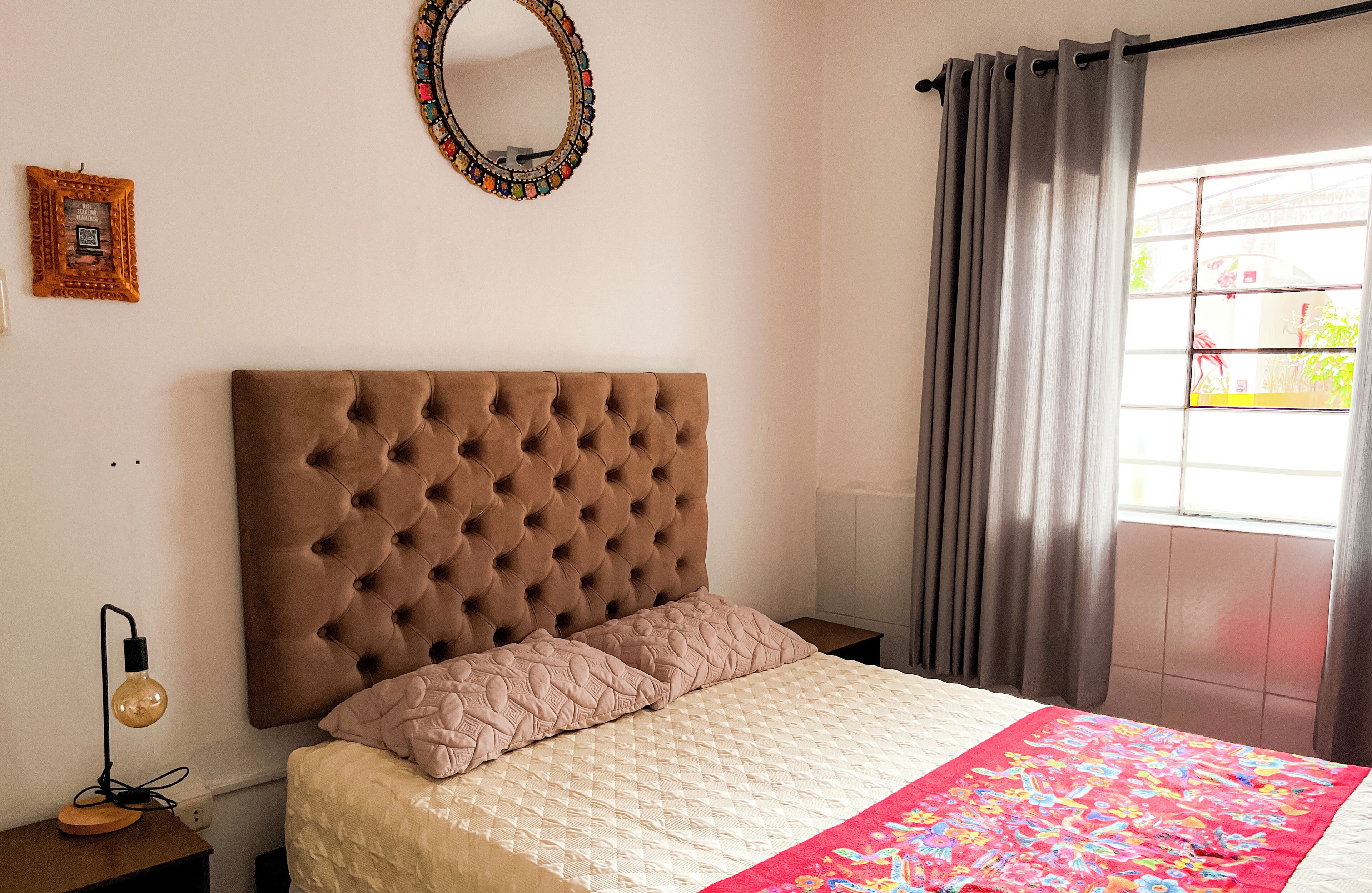 Un dormitorio con cabecera capitonada, cama con colcha acolchada, lámpara de noche, espejo decorativo y ventana con cortinas.