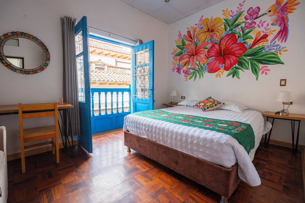 Ein Schlafzimmer mit Holzboden, einem Bett mit weißer Steudecke, einer blauen Tür und einem farbenfrohen floralen Wandgemälde.