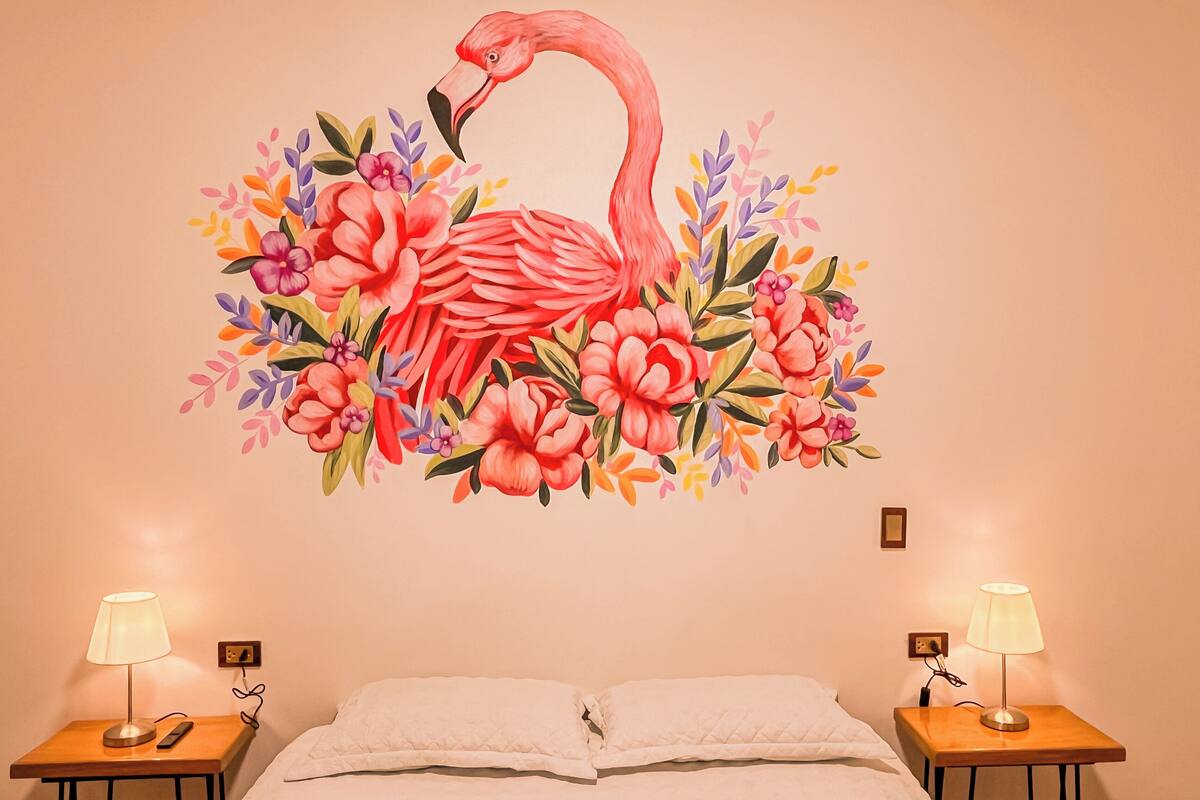 Ein Schlafzimmer mit einem Bett, zwei Nachttischen mit Lampen und einer Wandmalerei einer Flamingos, umgeben von Blumen.