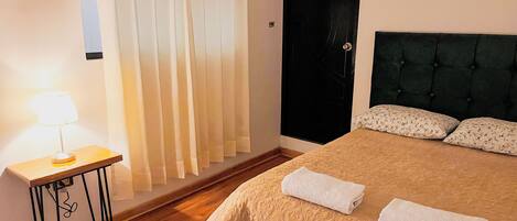 A neatly made bed with two towels, a bedside table with a lamp, a dark headboard, and a window with curtains.