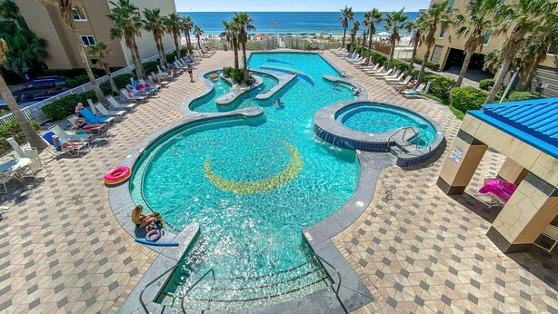 A swimming pool with multiple sections and a slide, surrounded by lounge chairs and palm trees, with a view of the ocean. - GULF VIEW, POOLS, HOT TUBS, BEACH ACCESS! (Gulf Shores)