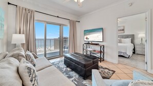 A spacious living room with a large window, a ceiling fan, a black ottoman, and a bed in the adjacent room. - GULF VIEW, POOLS, HOT TUBS, BEACH ACCESS! (Gulf Shores)
