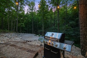 An outdoor grill with four burners and a stainless steel finish, set against a backdrop of a forested area with trees and a gravel path.