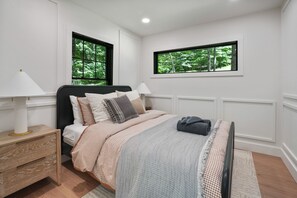A bedroom with a bed, bedside tables, and two windows with a view of trees.