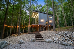 A modern cabin with a stone staircase leading to a wooden deck, surrounded by trees and a gravel path. - Uckor's Edge | Hocking Hills, Modern Cozy Cabin (Rockbridge)