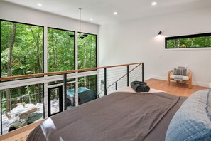 A bedroom with a large bed, a view of a forest through large windows, a small balcony, and a wooden chair.