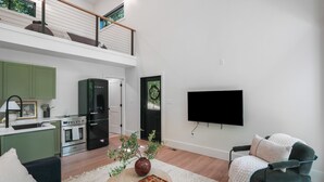 A modern living room with a flat-screen TV, a sofa, a coffee table with a plant, and a kitchen area with green cabinets and appliances. - Uckor's Edge | Hocking Hills, Modern Cozy Cabin (Rockbridge)