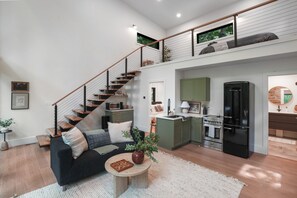 A modern loft-style apartment with a staircase leading to a loft bedroom, a kitchen with a black refrigerator, and a living area with a black sofa and a small round table.
