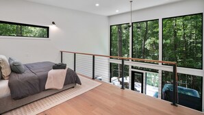 A modern bedroom with a large window, a balcony, and a view of a forest. - Uckor's Edge | Hocking Hills, Modern Cozy Cabin (Rockbridge)