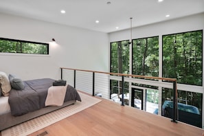 A modern bedroom with a large window, a balcony, and a view of a forest. - Uckor's Edge | Hocking Hills, Modern Cozy Cabin (Rockbridge)