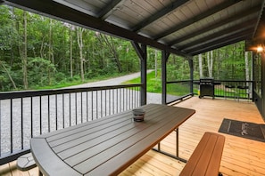 Un terrazzo in legno all'aperto con una panchina e un barbecue, circondato da una zona boschiva.