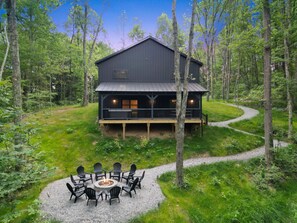 Une cabane dotée d’une terrasse, entourée d’une forêt, et un foyer avec des chaises.