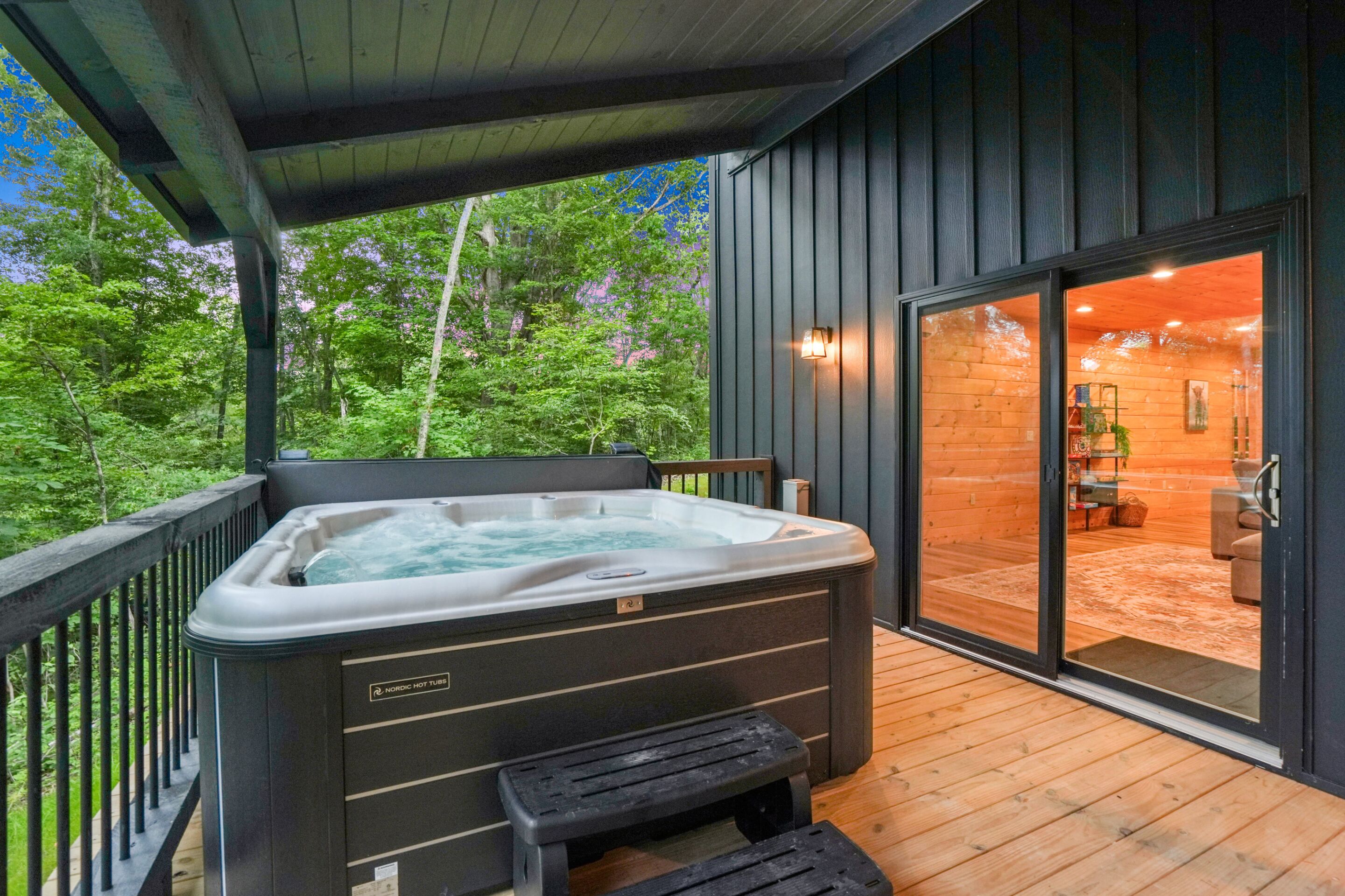 A hot tub on a wooden deck with a view of a forest and a building with a glass door.
