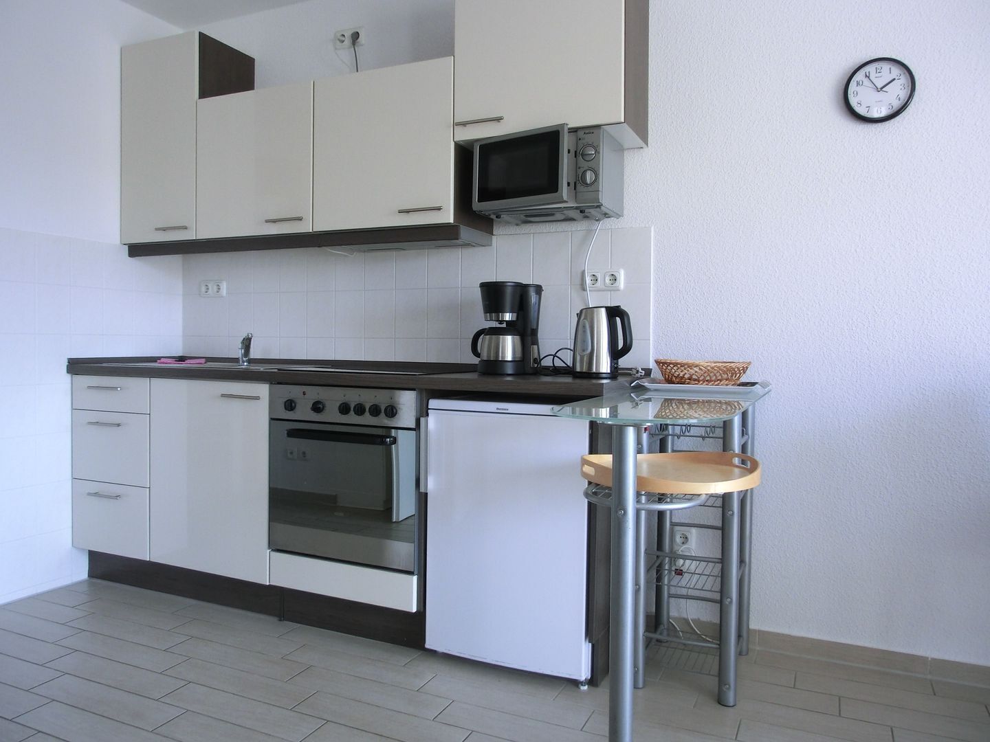 A compact kitchen with white cabinets, a microwave, an oven, a coffee maker, and a small table with a basket.