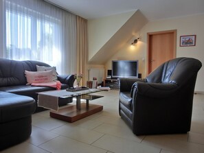 A living room with a black leather sofa, a glass coffee table, and a black leather armchair. A flat-screen TV is on a stand, and there's a door with a framed picture on the wall.