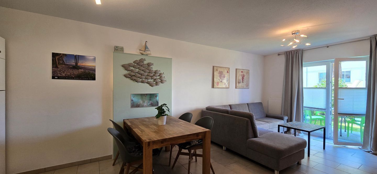 Ein modernes Wohnzimmer mit einem Holzesstisch, einer grauen Couch und einem kleinen Beistelltisch. An der Wand befinden sich gerahmte Bilder und ein Segelboot als Dekoration.