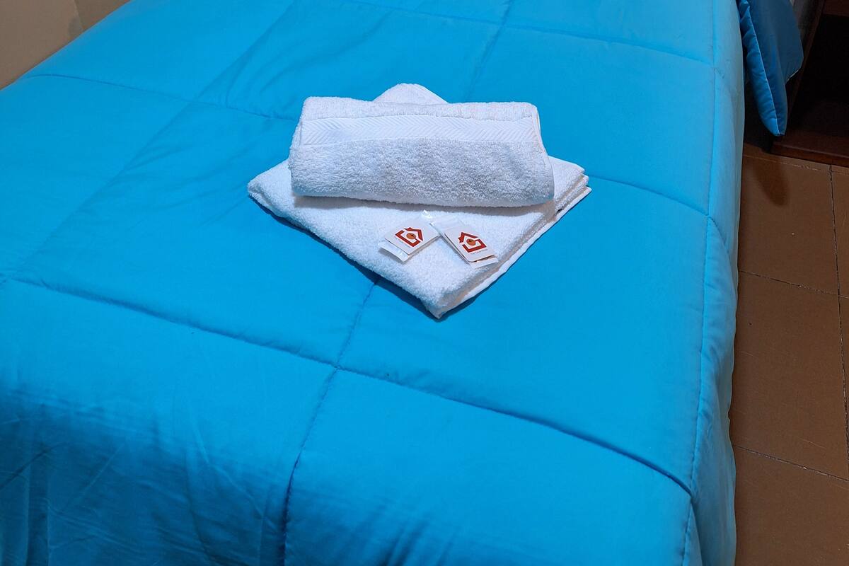 A neatly made bed with blue bedding and a folded white towel with a logo.