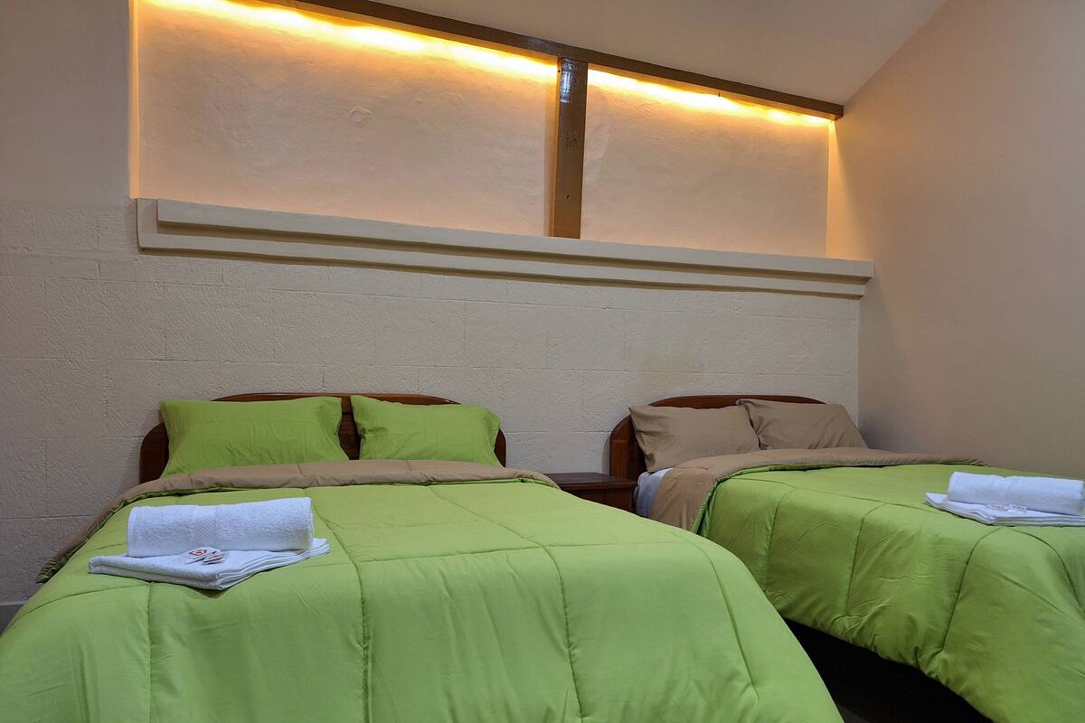 A hotel room with two beds, each with green bedding and a white towel. There is a wooden headboard and a wall-mounted light fixture above the beds.