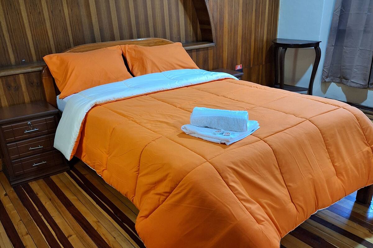 A bed with an orange bedspread and pillows, a wooden headboard, a small bedside table, and a folded towel on the bed.