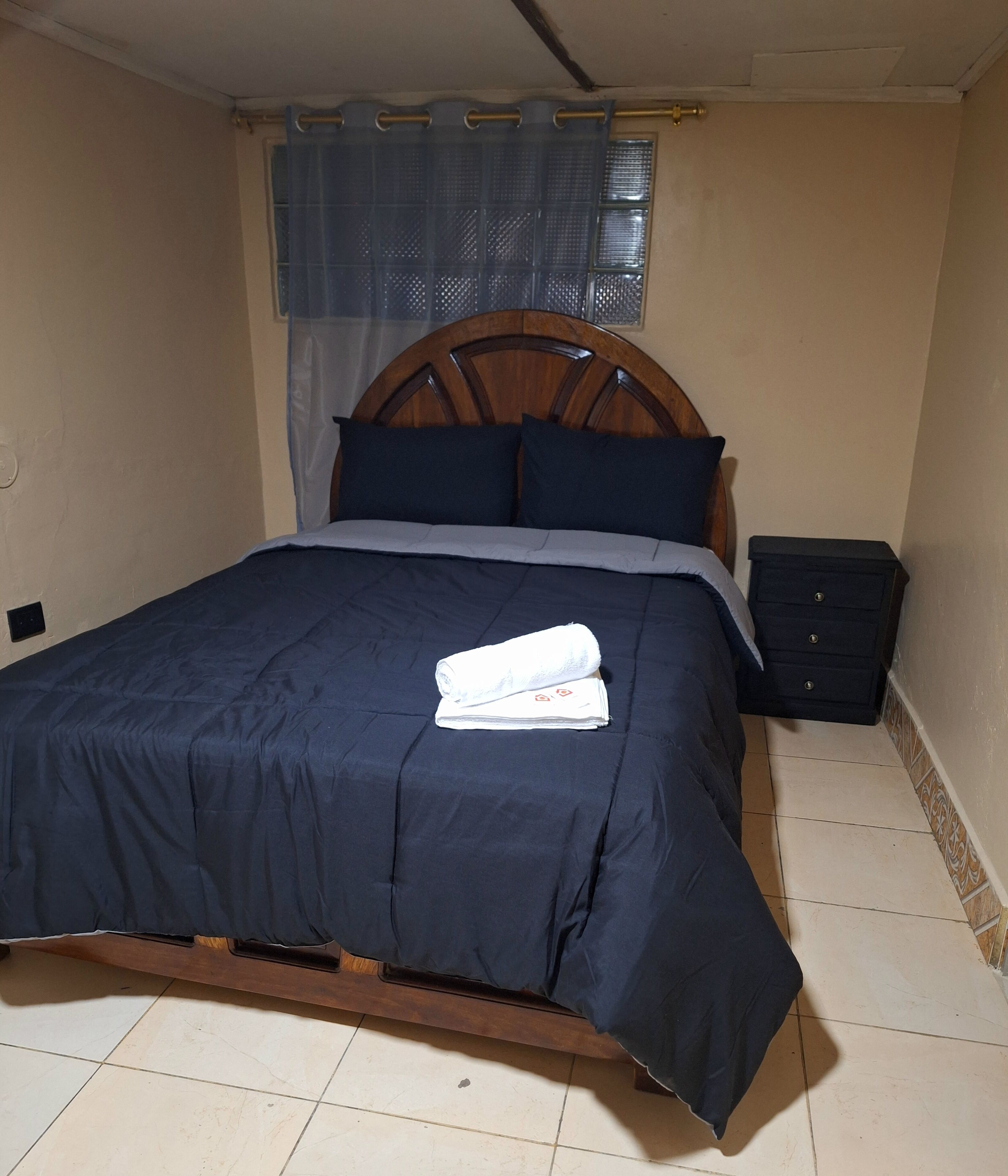 Un lit bien fait, recouvert d’une couvre-lit bleu foncé et agrémenté d’oreillers, avec une tête de lit en bois, une petite table de chevet dotée de tiroirs et une fenêtre ornée d’un rideau bleu.