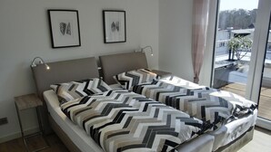 A double bed with a geometric patterned bedspread, a bedside table with a lamp, and two framed artworks on the wall. - Ferienwohnung Lp24 - Strandnahe 2-zi.-fewo für 2 Pers.- im 4. OG in Ruhiger Lage (Graal-Müritz)