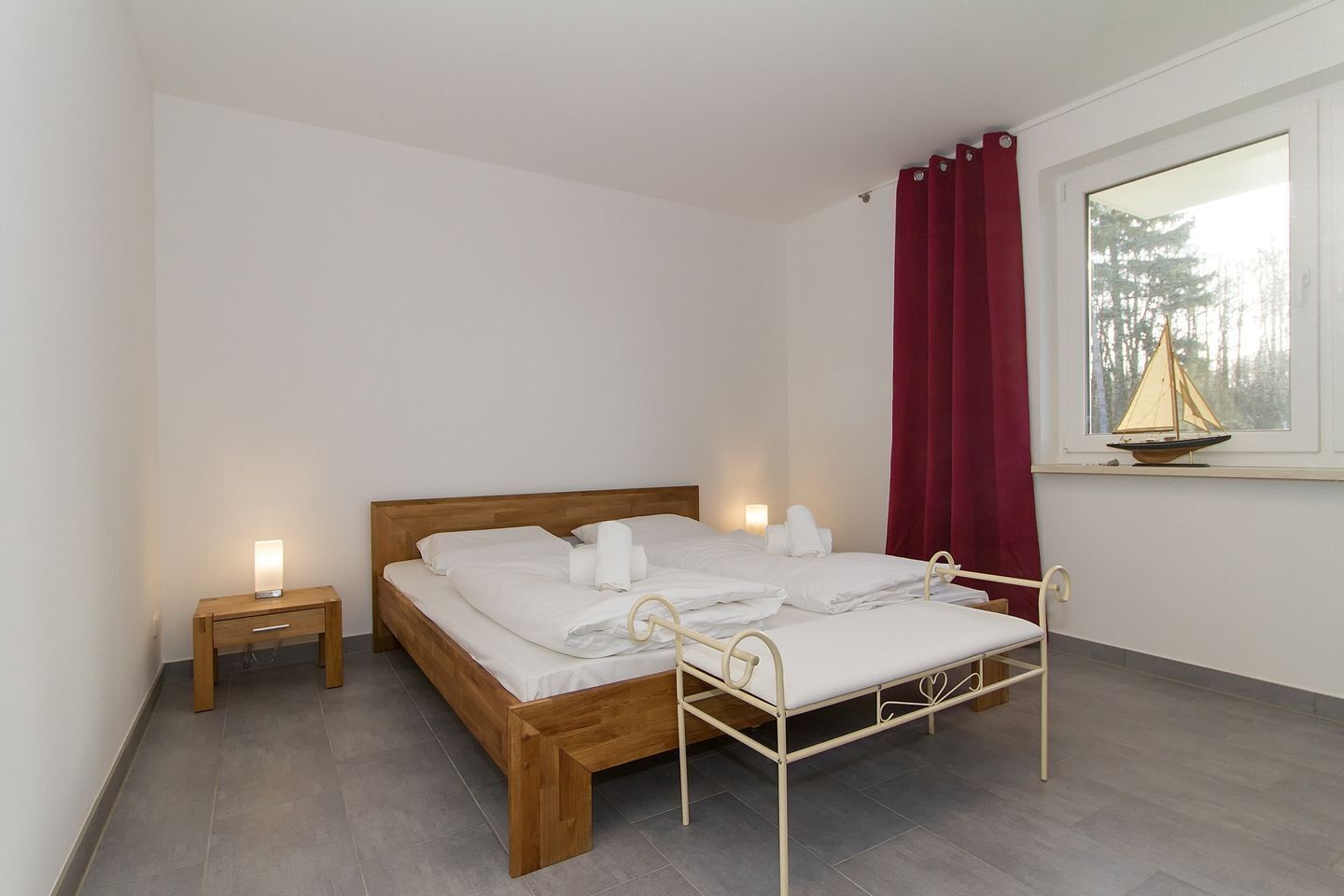 En pænt redt seng med hvide sengetæpper, et træsengegærde og en metalramme. Der er et lille natbord med en lampe, og et vindue med gardin og en modelsejlbåd på vindueskarmen.