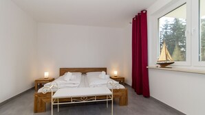 Un letto matrimoniale con lenzuola bianche, due comodini con lampade, una finestra con tende rosse e una piccola decorazione a forma di vela da barca sul davanzale.