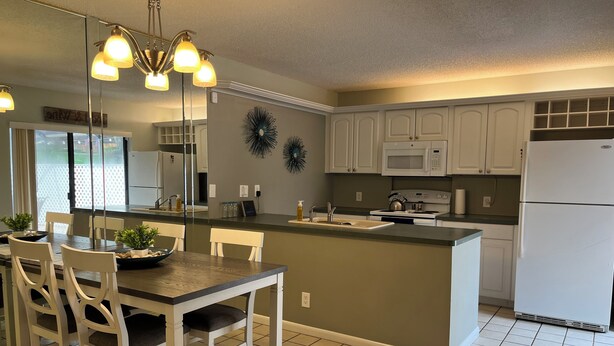A modern kitchen with white cabinets, a central island, a dining area with chairs, and a chandelier.