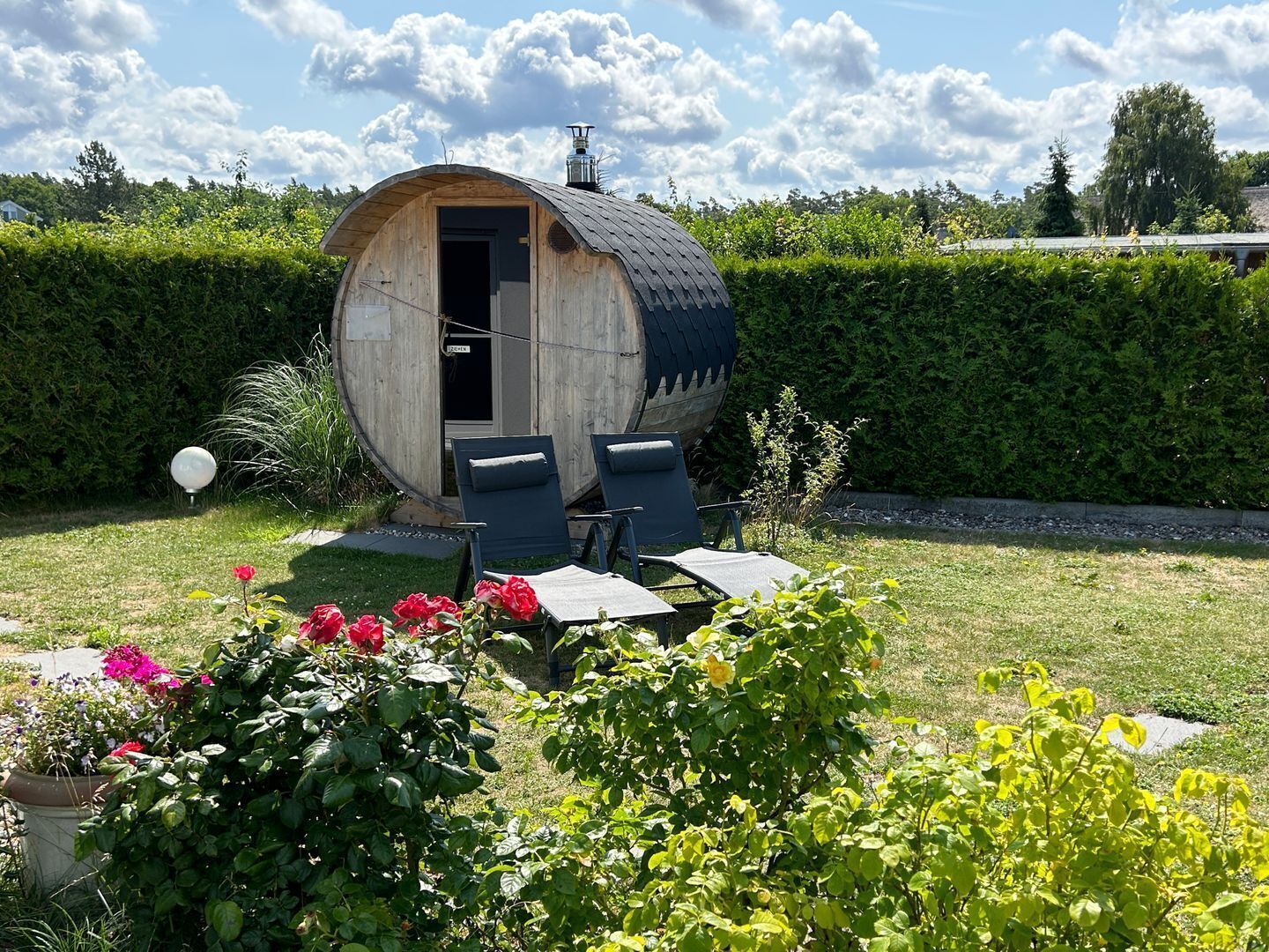 Eine rustikale Holzbütte mit Tür, zwei blauen Stühlen und einem kleinen Tisch in einem Garten mit verschiedenen Pflanzen und einem Hecken im Hintergrund.