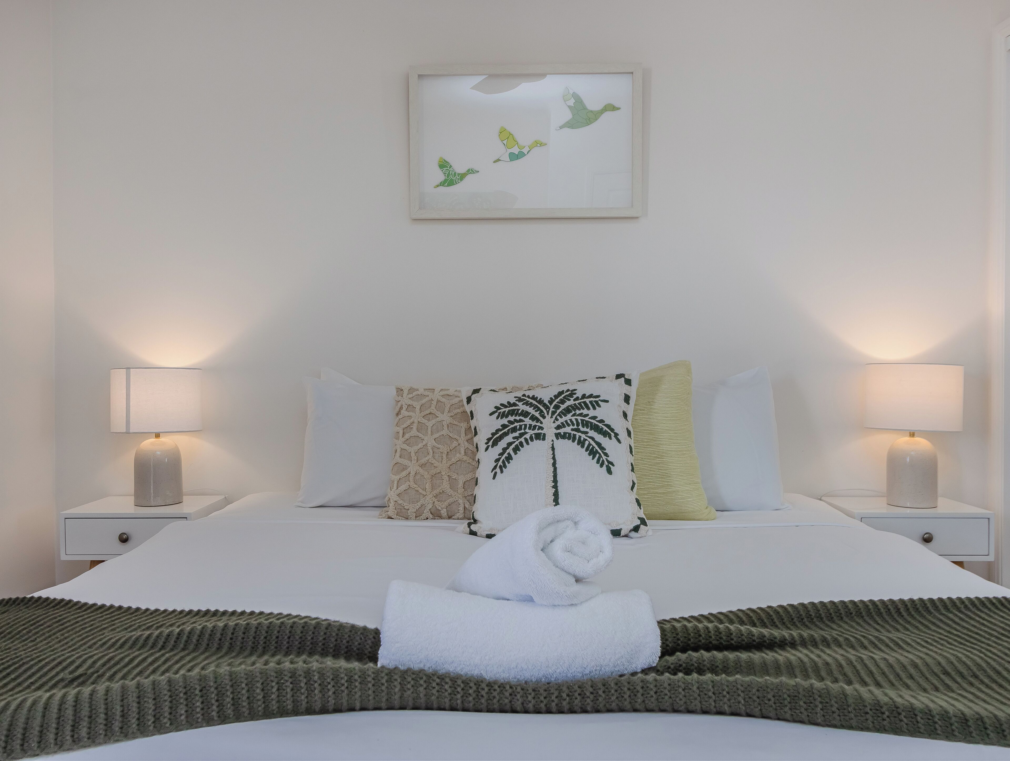 A neatly made bed with a folded towel, two bedside lamps, and a framed artwork of birds on the wall.