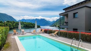 A rectangular pool with clear blue water, surrounded by lounge chairs and umbrellas, with a mountain backdrop and a building to the right. - Lake's Dream - MyHomeinComo (Ossuccio)