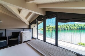 A bedroom with a bed, a dresser, two chairs, and a large window with a view of a lake and mountains.