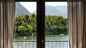 A view through a window with curtains, featuring a river, boats, and forested mountains. - Lake's Dream - MyHomeinComo (Ossuccio)