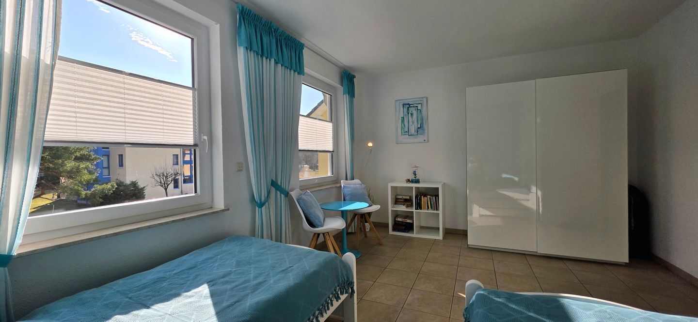 A small, well-lit room with two beds, a desk, a chair, and a wardrobe.