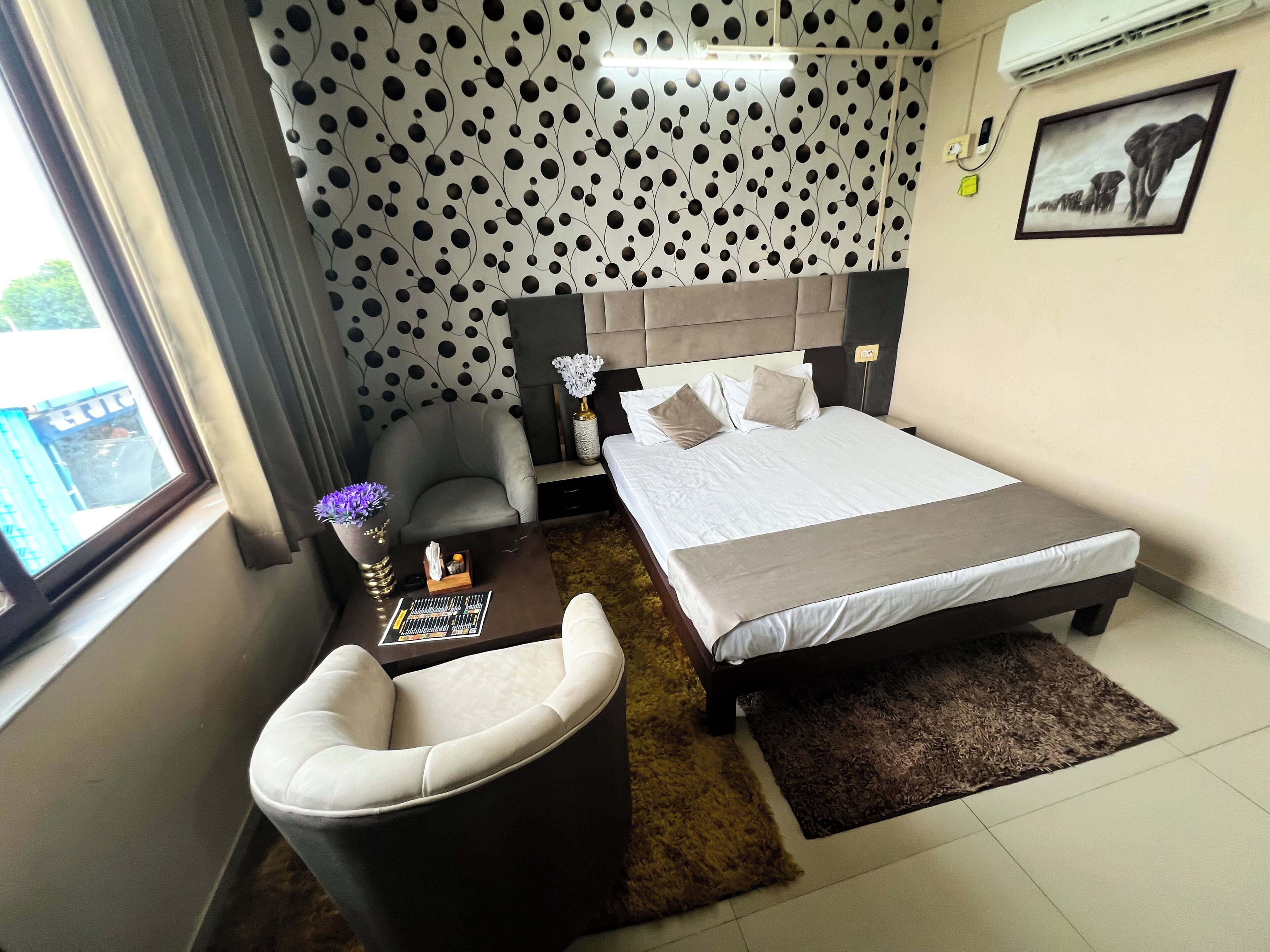 A hotel room with a bed, a chair, a small table with a vase, and a patterned wall.