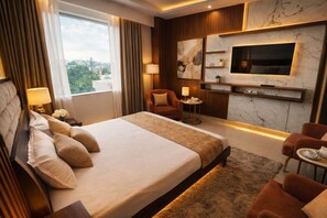 Habitación de hotel con una cama grande, televisión montada en la pared, zona de estar con una silla y una mesa, y vista al exterior.