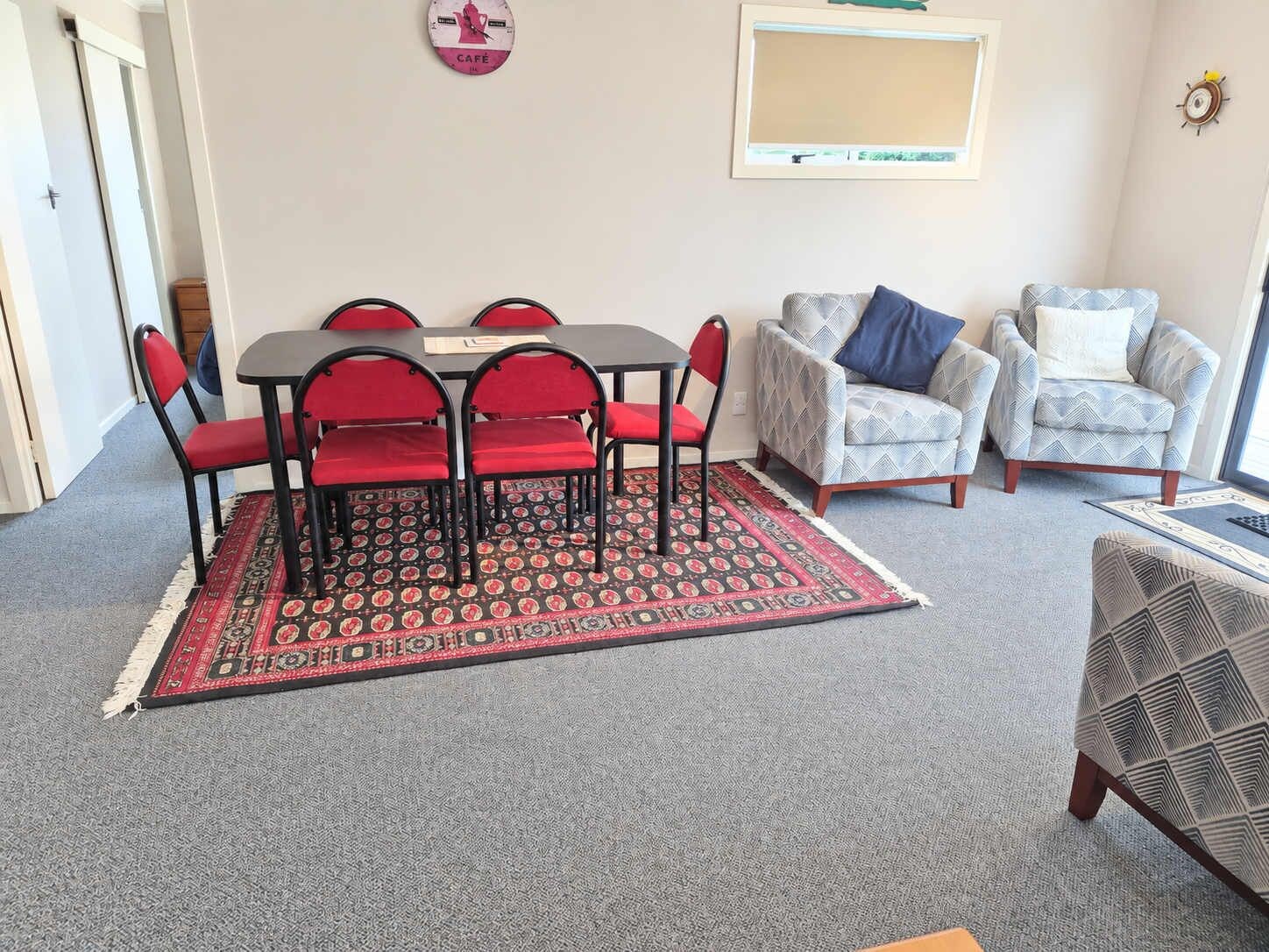 A room with a dining table and chairs, two armchairs, a patterned rug, and a clock on the wall.