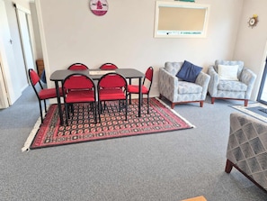 Ein Zimmer mit Esstisch und Stühlen, zwei Sesseln, einem gemusterten Teppich und einer Wanduhr.